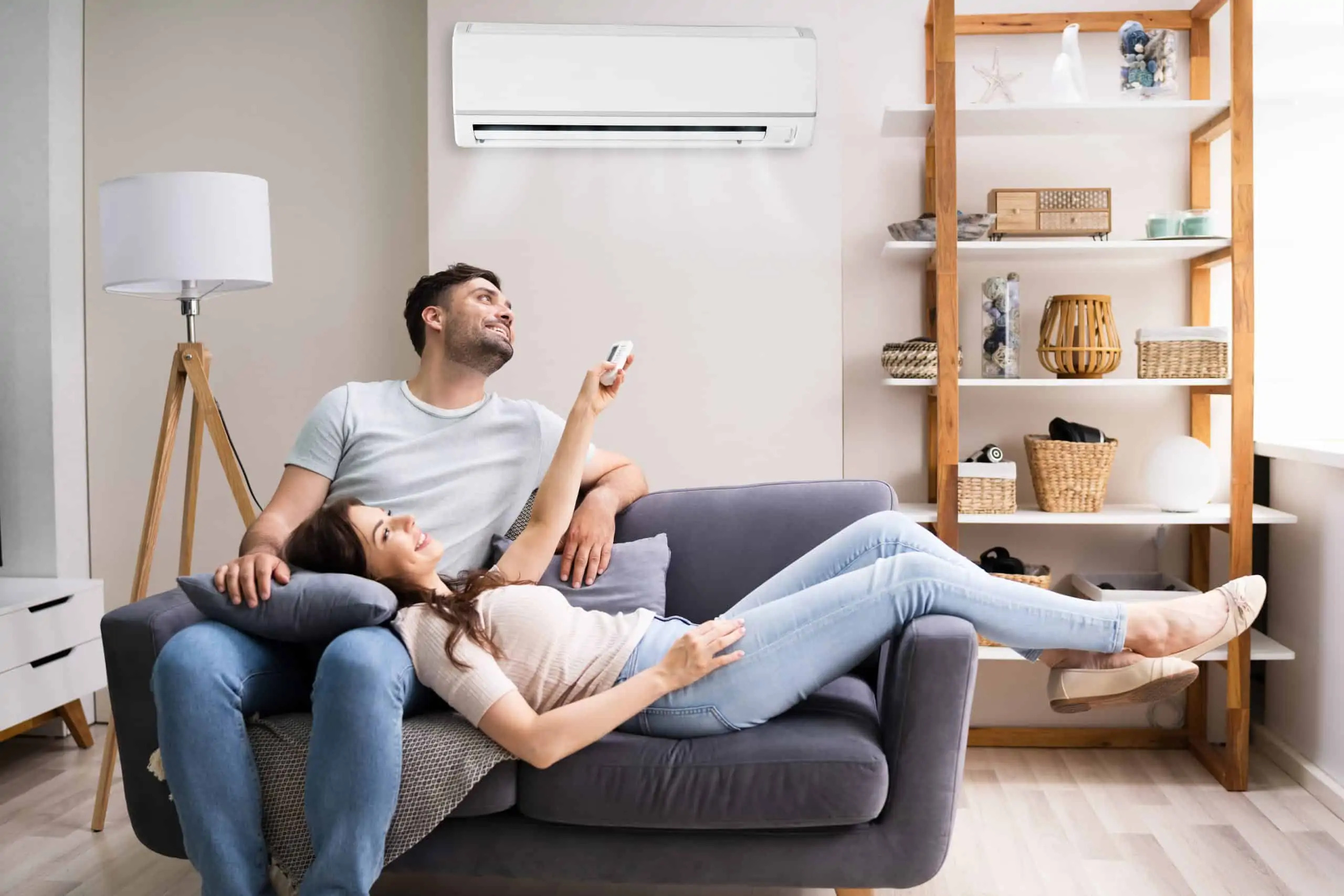 A couple relaxes on a gray couch in a modern living room with light-colored walls. The woman is holding a remote control and pointing it at a ductless air conditioning unit mounted on the wall above them.