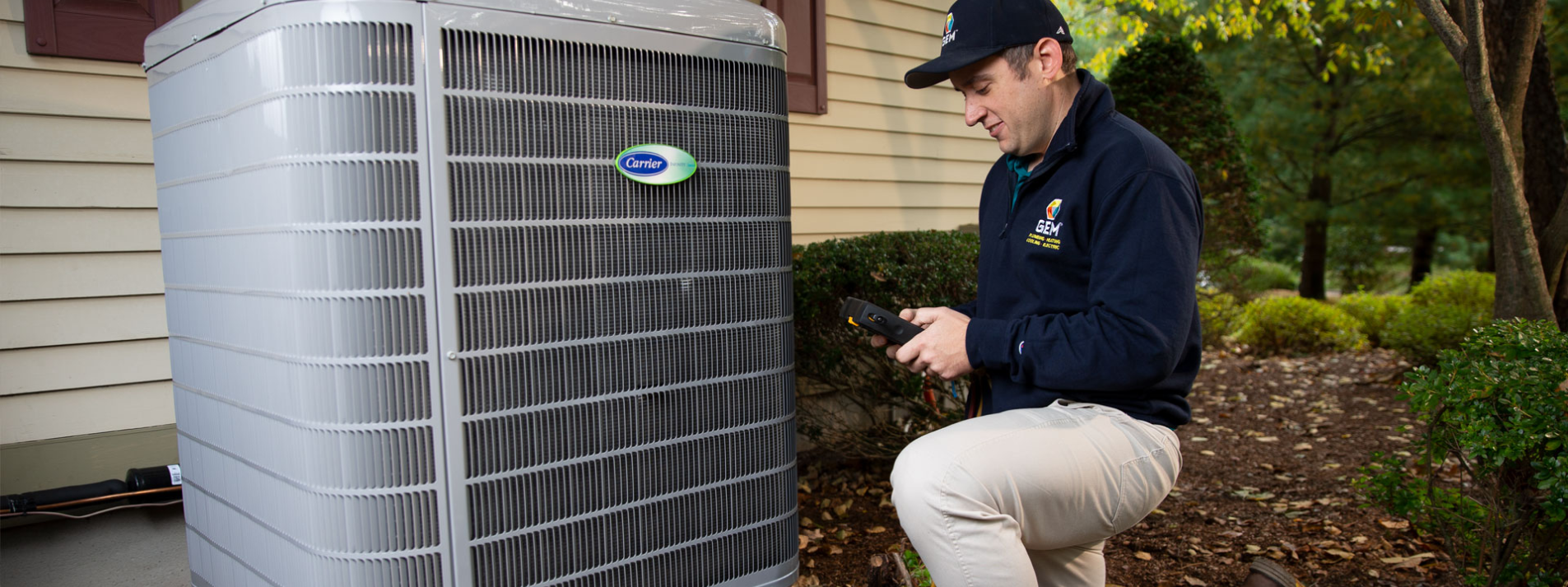 GEM technician performing service on Carrier HVAC unit near a house, surrounded by greenery.