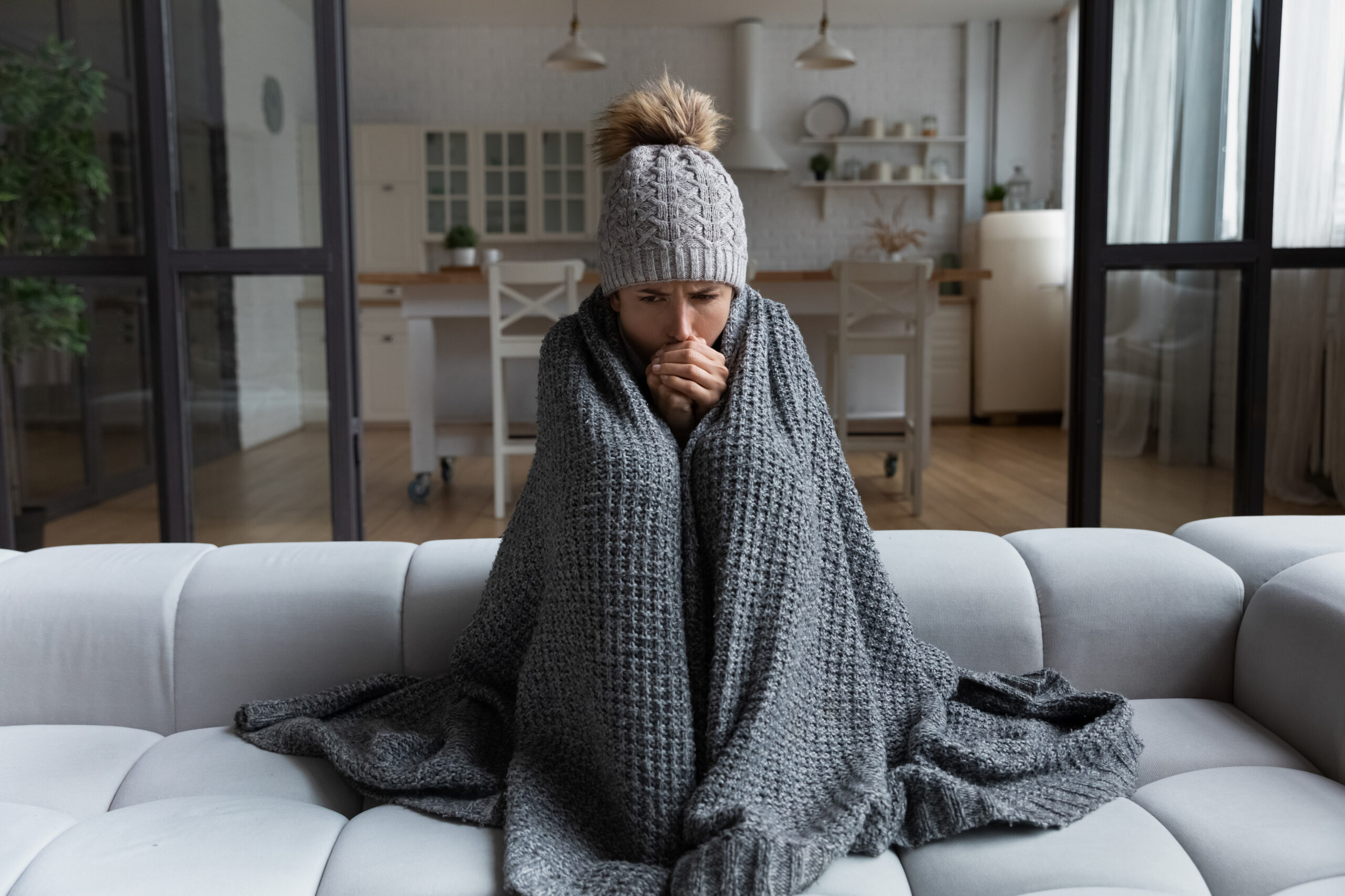 Woman sitting on couch in winter hat and blanket, shivering.