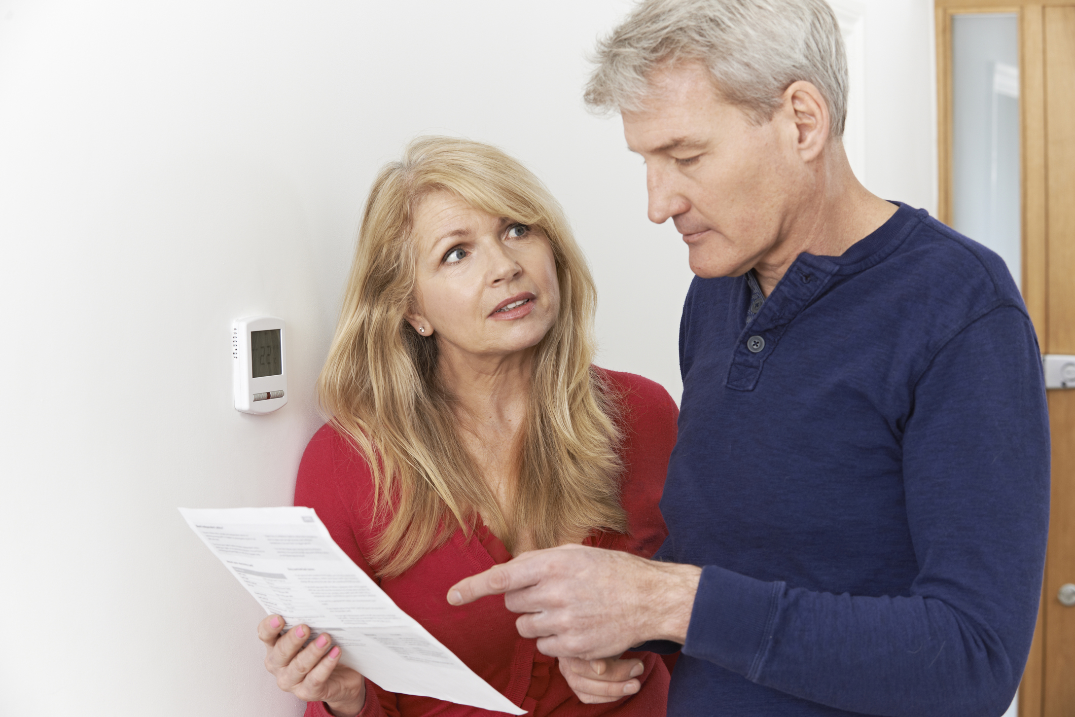 Worried Mature Couple With Bill, standing next to thermostat.
