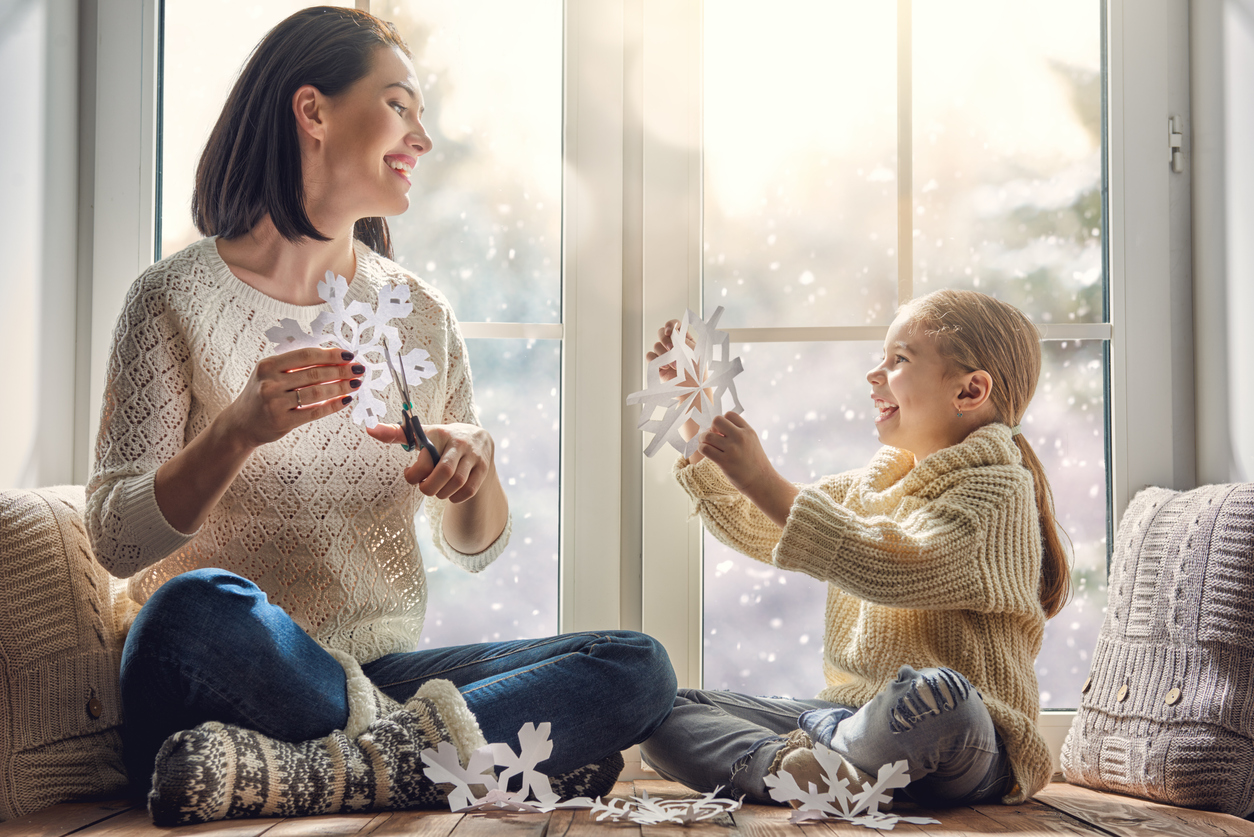Mother and young daughter making paper snowflakes in front of a window; snowy scene outside.