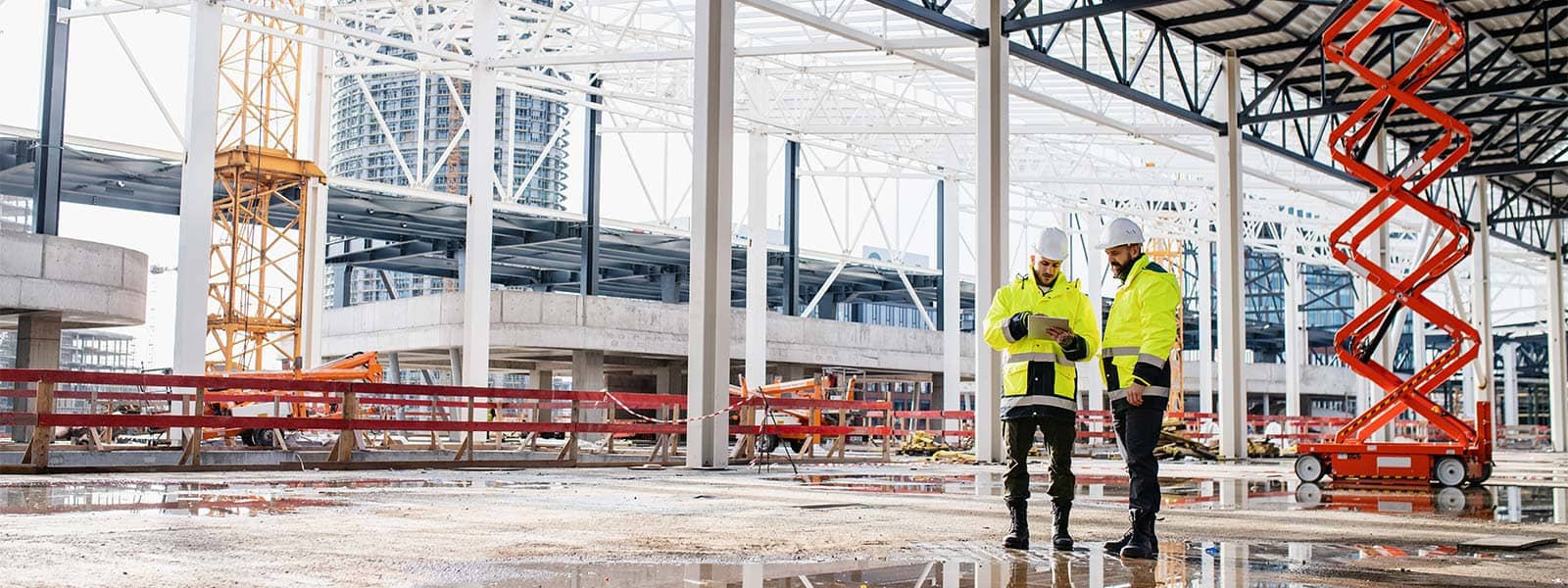 2 construction workers in Hi-Vis jackets and hard hats looking at a paper while standing in a building under construction.